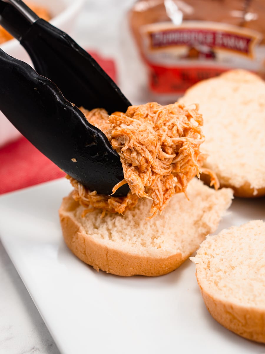 using tongs to place buffalo chicken on a slider bun