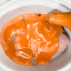 pouring buffalo sauce over chicken breasts in the slow cooker