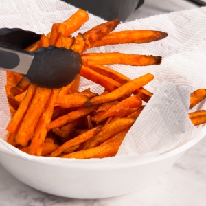 using tongs to move sweet potato fries to a paper towel lined bowl after cooking