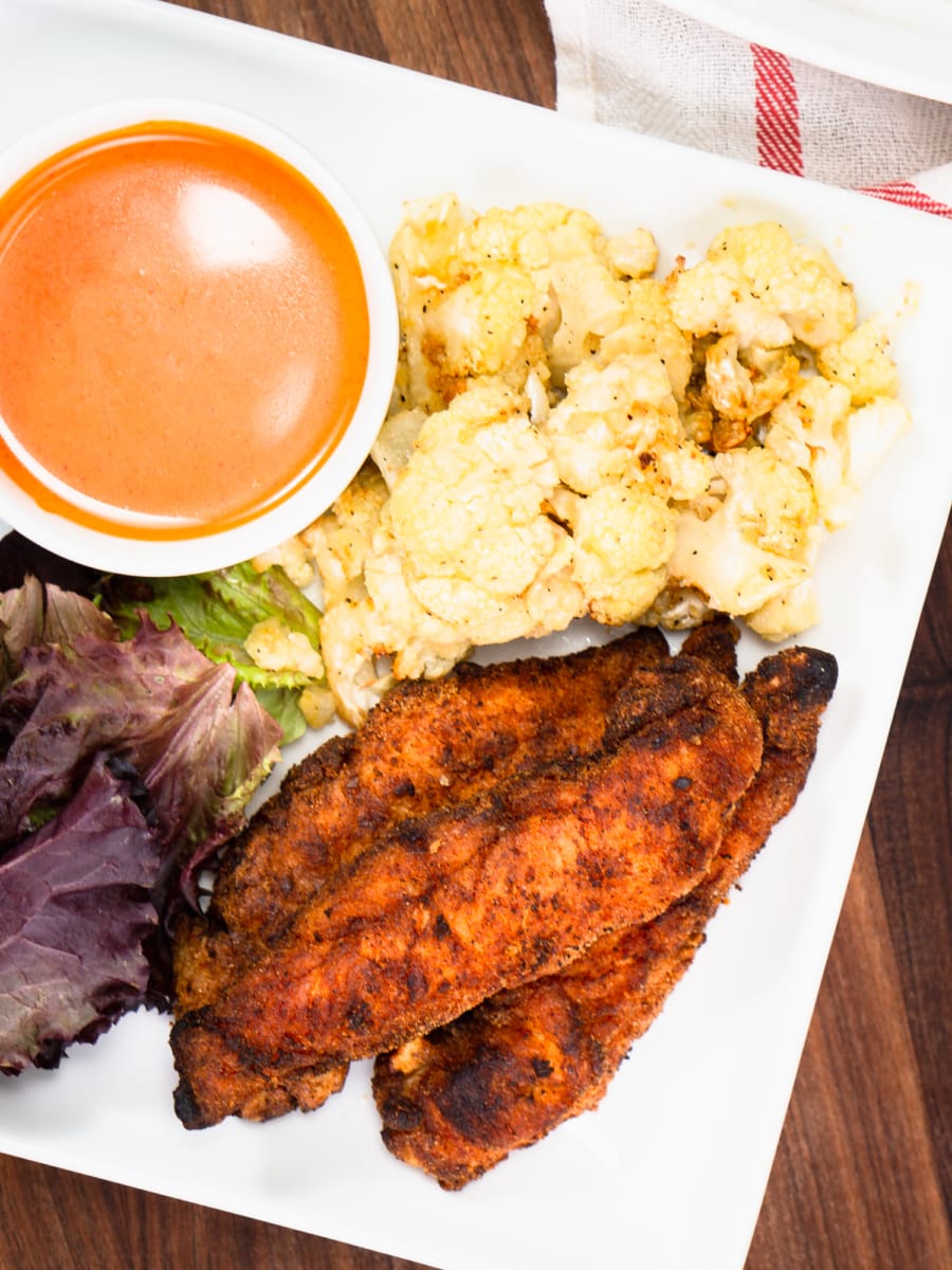 gluten free chicken tenders on a plate with gluten free buffalo sauce, cauliflower, and a salad