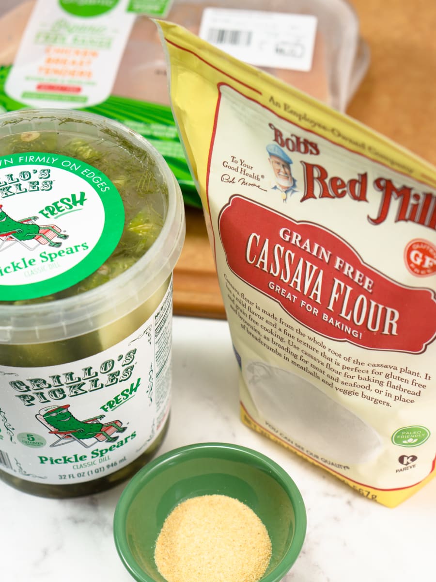 ingredients for gluten free chicken tenders including cassava flour, a jar of dill pickles, pasture raised chicken tenders, and garlic powder