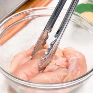 using tongs to add chicken tenders to a glass mixing bowl