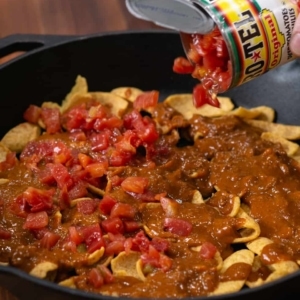 adding a can of rotel tomatoes and green chiles evenly over a layer of fritos and chili in a skillet to make frito chili pie